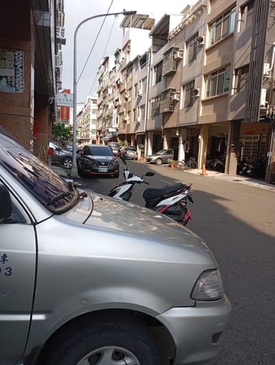 A sunny street scene in an urban area. The camera is at street level, focusing primarily on the front portion of a silver vehicle with blue text on the side. A newer model dark gray Nissan Kicks is parked behind the silver vehicle, further down the street. A small white motorcycle with red accents is parked on the right side of the street near the Nissan Kicks. In the background, the street stretches away from the camera, flanked by buildings, varying in height and architectural style, that appear to be residential or mixed-use buildings with multiple windows and air conditioning units. A streetlamp is visible on the left. The scene conveys an ordinary, bustling street in Kaohsiung, Taiwan.