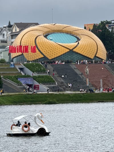 In Xuan Huong Lake, Da Lat, Vietnam, a swan-shaped pedal boat with a white canopy, carrying four people, navigates the choppy water. A lush green bank borders the lake, leading to a tiered landscape featuring grand staircases and terraced gardens. Dominating the background is a striking, petal-shaped building with a golden, tessellated roof and a central, turquoise pool. Red flags adorn the building's facade. Numerous people are visible on the staircases and surrounding pathways, engaged in various activities. The sky is overcast.