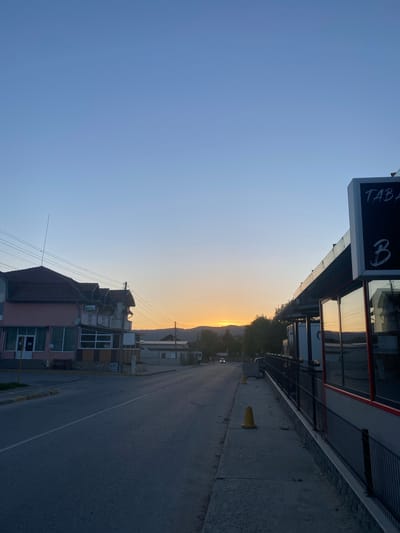 The image depicts a street scene in Satovcha, Bulgaria at sunset. The street runs between buildings, with the sky visible in the distance, exhibiting a gradient from blue to orange. On the left is a building with a sign that reads "TABA" followed by a Cyrillic letter. On the right is a building with reflective windows, a metal railing, and a concrete sidewalk. Traffic cones are positioned along the sidewalk. A street runs between these buildings. Buildings can be seen in the distance. Overhead power lines are visible.