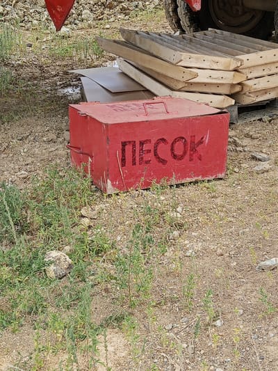A weathered red metal box with the Cyrillic word "ПЕСОК" (meaning "sand") painted in black on its side sits on gravelly ground. The box has a single metal handle on its lid. Behind the box, a stack of wooden planks is visible. The scene appears to be outdoors, with sparse green vegetation growing around the box and on the ground. In the background, a portion of a large vehicle tire and a red triangular object are partially visible. The overall environment suggests a construction or storage area, possibly in a rural or semi-industrial setting. The specific location in Raygorodok, Kazakhstan, is not visually identifiable from the image itself.