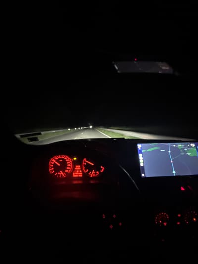 The image shows the interior of a car at night. The dashboard is visible with illuminated red gauges, including a speedometer and tachometer. A navigation screen displays a map. Streetlights are visible through the car's side window. The location, Mogila, Bulgaria, cannot be determined from the image. No people are visible.