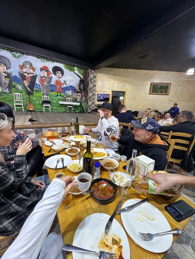 A group of people are gathered around a wooden table laden with food and beverages in what appears to be a restaurant. The table holds plates, bowls, glasses, silverware, and bottles, suggesting a meal is in progress. Several individuals are visible around the table, some seated on wooden chairs. One person is wearing a cap with "Blues" visible on it. Another person wears a black cap and a white t-shirt.
In the background, a wall mural depicts figures in historical or traditional attire. The restaurant's interior features stone-like wall patterns and dim lighting. A painting hangs on the wall above the people in the background. Based on the prompt, this scene could be taking place in Kveda Chkhutuneti, Georgia.