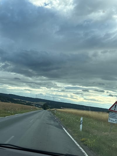 The image depicts a road in Oberwetz, Germany, viewed from a car. The sky is partly cloudy, with a mix of dark, dramatic clouds and brighter, sunlit patches, suggesting a dynamic weather pattern. The time of day appears to be late afternoon or early evening, given the low angle of the sun.
The scene shows a two-lane asphalt road stretching into the distance, bordered by grassy fields and a treeline on the right. On the left side of the road, there are several wind turbines visible in the middle distance, indicating a rural or semi-rural landscape. A white road marker with black markings is present in the field to the right of the road.
Attached to a post on the right side of the road are two German traffic signs. The top sign is a triangular warning sign with a red border and an exclamation mark, indicating a general hazard. Below it, a rectangular sign has text in German and an arrow pointing upwards. The text reads "Straßen- schaden" and "1,8 km". This translates to "Road damage" and indicates that the road damage is 1.8 kilometers ahead.
The overall mood is somewhat dramatic due to the contrast between the dark clouds and the sunlight. The image captures a moment of travel on a German road, with visual cues of both natural elements and man-made infrastructure.