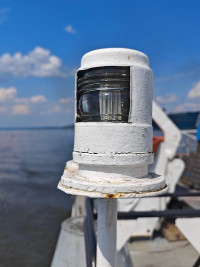 A close-up, eye-level, outdoor shot features a white, weathered navigation light on a boat. The light, a prominent cylindrical object with a glass lens and ribbed detailing, is centrally positioned and appears to be in use, though not illuminated. The paint on the light is chipped and shows signs of rust. In the background, a body of calm water stretches towards a hazy horizon, with a faint shoreline visible in the distance. The sky above is a vibrant blue, dotted with scattered white clouds. The boat's railing and other structures are visible to the right, partially out of focus. The scene evokes a sense of maritime activity under a clear, sunny sky. The image does not contain any individuals.