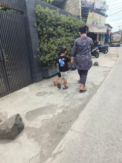 A woman and two children walk along a sidewalk. The woman wears a patterned gray outfit and sandals, carrying a plastic bag. One child, in a black shirt and patterned shorts, walks beside her, while another smaller child follows behind. They are walking past a fence with greenery and a building. In the background, motorcycles are parked near a building with a sign, and a truck is visible further down the street.