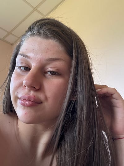 This media file is a close-up, portrait-style photograph of a young woman, likely indoors. The image is oriented sideways. The woman has long, dark brown hair that is falling around her face. Her eyes are brown, and she has dark eyebrows. Her lips appear to be slightly parted and are a muted pink color. There are a few small blemishes visible on her skin, particularly on her cheeks and chin.
The background appears to be a plain, light-colored ceiling with some grid-like patterns. The lighting suggests it is daytime, but no specific external weather conditions are visible. The woman's expression is neutral, with a slight hint of a smile. A hand with white-painted fingernails is visible in the upper portion of the frame, gently touching her hair. A thin red string or bracelet is also visible on her wrist, peeking out from her hair.
The overall impression is a casual, candid selfie taken in what appears to be a domestic setting. No text is visible in the image.
