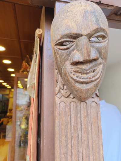 A carved wooden mask with an exaggerated smile and hollow eyes is mounted on a wooden post. The mask is weathered and shows signs of age. In the background, a shop interior is visible with shelves displaying various items, illuminated by overhead lights. A partially visible person wearing a white shirt stands to the right of the mask. The scene appears to be an outdoor setting, possibly a market stall or a shop entrance in Gokarneshwar, Nepal.