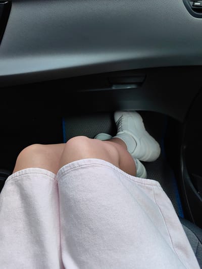 The image shows a close-up view of a person's legs inside a vehicle. The person is wearing a light pink skirt and white high-top sneakers. The background consists of the car's interior, including the dashboard and a floor mat. The lighting suggests it is daytime, and the scene appears to be captured from a seated position within the vehicle. The location of Хаценчыцкі сельскі Савет, Belarus is not depicted in this image.