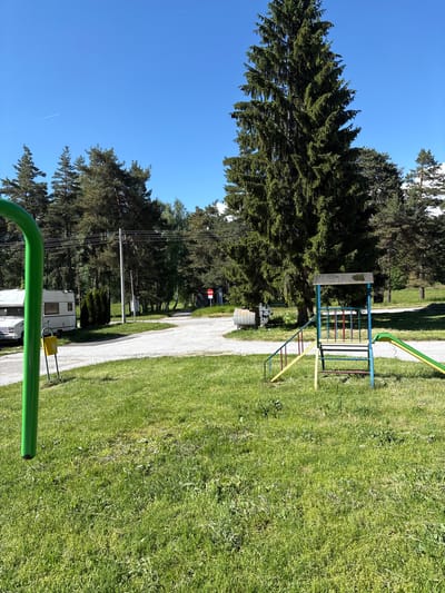 The image depicts a grassy park area in Panagyurski kolonii, Bulgaria, under a clear blue sky. A large evergreen tree stands prominently on the left. A gravel path runs through the center of the image, leading into the distance. A colorful playground structure, with blue, yellow, and red bars, is visible on the right side of the path. Further down the path, a white camper is parked. In the foreground, a curved green object is partially visible. Power lines run along the left side of the path. A no-entry sign is situated further along the path.