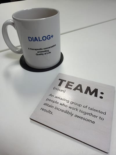 A white mug with the inscription "DIALOG+ A therapeutic conversation promoting Quality of Life" rests on a black coaster on a white surface. Next to the mug, a white card or coaster with the text "TEAM: (noun) An amazing group of talented people who work together to attain incredibly awesome results." is placed. The scene is likely indoors and situated on a desk or table. The location in the city of Mold, United Kingdom is unknown.