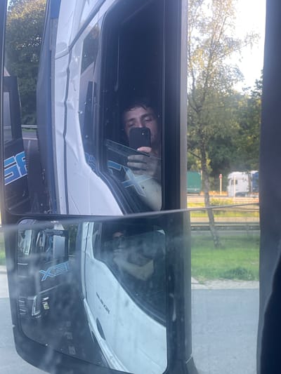 This image captures a close-up reflection in a vehicle's side mirror, likely a truck given the perspective. The primary subject is a person's face, partially visible and appearing to hold a smartphone, suggesting they are taking a selfie or using the phone. The reflection shows a person with dark, curly hair and a light complexion. The setting appears to be outdoors, with trees and greenery visible in the background, indicating a daytime scene. The lighting suggests it's a bright day, possibly late morning or early afternoon, with sunlight filtering through the leaves.
The reflection in the mirror also reveals parts of the vehicle's interior, including the dashboard and window frame. A white vehicle is reflected prominently in the main mirror, with some blue lettering that is partially legible as "ISX." The scene is likely taking place in Ganderkesee, Germany, as indicated by the provided location context. There are no overt signs of distress or specific emotions conveyed, other than the casual act of taking a photo. The overall impression is that of a brief moment captured during a journey or a stop, where the driver is interacting with their phone and observing their surroundings through the mirror. The image is oriented vertically, suggesting it might have been taken while the vehicle was parked or moving.