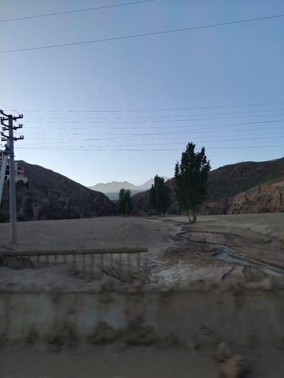 A wide shot captures a dry, sandy riverbed in Bulan-Segettu, Kyrgyzstan, under a pale, clear sky. The landscape is dominated by barren, rocky hills on either side, their slopes covered in sparse vegetation. In the foreground, a rudimentary wooden fence runs horizontally across the frame, partially obscuring a rough, sandy embankment. A weathered utility pole with multiple crossbars and power lines stands prominently on the left side of the image, with the skeletal remains of a building visible behind it. Further into the scene, several tall, slender trees grow along the riverbed, their dark green foliage stark against the muted tones of the landscape. A faint, shallow stream, marked by lighter, wet sand, meanders through the middle ground. In the distance, a range of hazy mountains forms a soft horizon line against the sky. The overall impression is one of a remote, arid, and undeveloped environment.