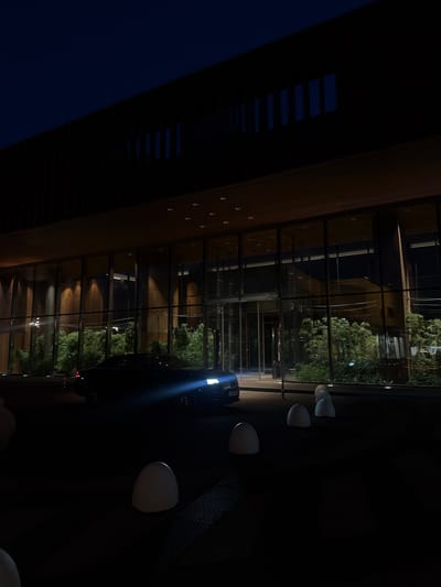 The image depicts an exterior view of a modern building at night, possibly a hotel or administrative complex, located in the city of Barvikha, Russia. The building features a dark, horizontal facade with a band of windows running across the lower level, illuminated from within by warm lighting that casts shadows on the interior wooden paneling. Above the windows, the facade is dark and featureless, with a series of vertical slits visible against the night sky.

In front of the building, a dark-colored luxury sedan is parked, with its headlights on, casting a bright beam onto the paved area. Several white, egg-shaped bollards are arranged in a line across the foreground, some of which appear to be illuminated. Behind the car and visible through the glass facade are dense green shrubs or trees, suggesting landscaping. The overall atmosphere is one of quiet elegance and sophistication, typical of upscale establishments.