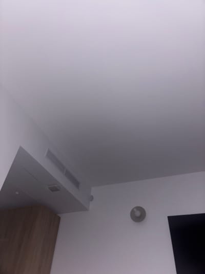 The image shows a corner of a room, likely indoors. The walls are painted in a light, neutral color, perhaps off-white or a very pale gray. A flat-screen television, partially visible, is mounted high on the wall, with only the upper corner appearing in the frame. Below the TV, a small, circular object, possibly a light fixture or a smoke detector, is affixed to the wall.
Beneath the light fixture and the television, there is a white cabinet with a streamlined, minimalist design. It has inset handles and a smooth surface. Adjacent to the cabinet, there appears to be the top surface of a wooden dresser or a desk, with a wood grain pattern that suggests it is made of laminate or veneered wood.
The scene is sparse and modern, with a focus on clean lines and simple geometric shapes. There is no indication of the location being Mellieħa, Malta, or any other specific geographic location.