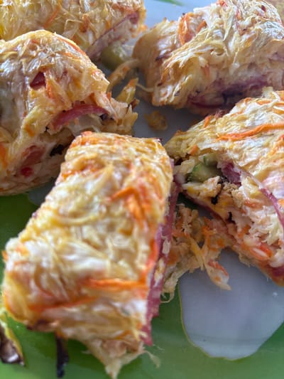 The image displays a close-up, overhead view of several pieces of cooked food, likely some sort of savory roll or strudel, arranged on a green and white patterned surface. The food items are cut into rectangular sections, revealing a filling that appears to contain shredded vegetables, possibly carrots, mixed with what looks like ham or salami and some creamy white substance. The outer layer is golden brown and appears crispy, with visible strands of shredded cheese or dough. The lighting is bright, casting some shadows. There is no indication of any specific location in the city of Shipovo, Russia, or any people present.