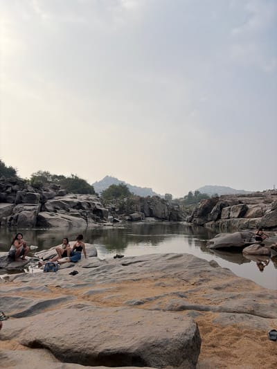 The scene depicts a natural river environment in Hale Bandi Harlapura, India, under an overcast or hazy sky. The foreground and midground consist of large, smooth, grey rock formations interspersed with patches of light brown sand. A calm body of water, likely a river, flows from the foreground towards the background, reflecting the surrounding rocks and sky.
Four individuals, appearing to be adult females, are present. On the left side of the midground, three individuals are positioned on the rocks near the water's edge. One is in a crouching posture, facing the water. The other two are seated; one is wearing a dark top and light-colored bottoms with a dark skirt or wrap, and the other is wearing a light top. A patterned blue and white bag is visible on the rock beside them, along with several pairs of black sandals.
Further into the midground, on the right side of the water, a fourth individual is seated on a rock near the water, partially obscured, with their reflection clearly visible in the water. This individual appears to be wearing dark swimwear or a dark top and bottoms.
The river is flanked on both sides by extensive formations of large, grey boulders and rocks, with sparse green vegetation and trees visible on and between the rock formations, particularly on the left bank. Distant hills or mountains are discernible in the far background beneath the pale sky. The overall impression is a serene, rugged landscape.
