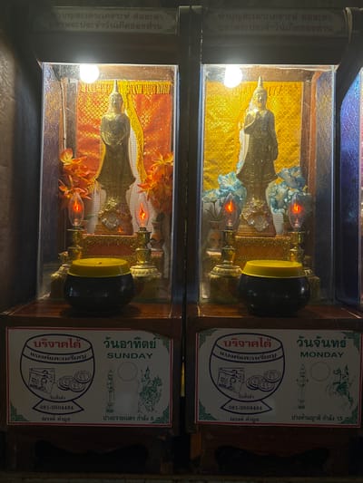 The scene depicts an interior view within a religious establishment in Ban Hin Sam Kon, Thailand. Two illuminated, glass-fronted wooden display cases stand side-by-side, each dedicated to a specific day of the week for offerings.
The left display case, associated with "SUNDAY," contains a standing, gold-colored Buddha statue with a pointed finial. The statue's right hand is raised in a gesture of blessing, and it is positioned against a textured, reddish-orange patterned fabric background. Artificial orange flowers flank the statue. Two flame-shaped electric candles emit an orange glow from the base, supplemented by a white light bulb above. Below this case, a black offering bowl with a yellow lid rests on a wooden shelf. A white sign with green borders identifies the day as "วันอาทิตย์" (Sunday) and "SUNDAY," displaying illustrations of banknotes and coins, a phone number (081-0504464), and a depiction of the Buddha posture "ปางถวายเนตร กำลัง 6" (Pang Thawai Net, strength 6).
The right display case, associated with "MONDAY," is similar in structure. It houses another standing, gold-colored Buddha statue, also with a pointed finial and its right hand raised, set against a textured, golden patterned fabric background. Artificial blue flowers flank this statue. Two flame-shaped electric candles provide an orange glow from the base, with a white light bulb illuminating from above. Below this case, an identical black offering bowl with a yellow lid sits on the wooden shelf. Its corresponding white sign with green borders identifies the day as "วันจันทร์" (Monday) and "MONDAY," also showing illustrations of banknotes and coins, the same phone number (081-0504464), and a depiction of the Buddha posture "ปางห้ามญาติ กำลัง 15" (Pang Ham Yat, strength 15).
Both displays are integrated into dark wooden cabinetry. Partially visible Thai script adorns the top section of the structure above the cases, likely indicating spiritual dedications or instructions. No individuals are present in the scene. The lighting is localized, highlighting the religious artifacts and offering stations.