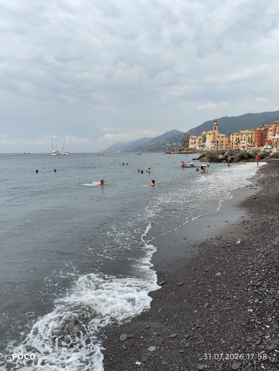 The image depicts a view of the Camogli waterfront and beach in Italy. In the foreground, a dark, pebble-strewn beach meets the choppy gray sea, with waves creating white foam as they wash ashore. Several people are in the water, swimming or wading. In the middle ground, sailboats are visible on the sea. The background features the colorful buildings of Camogli, characterized by their narrow facades and red-tiled roofs, clustered along the coastline. Behind the town, lush green hills rise steeply. The sky is overcast with gray clouds. The lower left corner of the image shows the text "POCO F5," and the lower right corner displays the date and time "31.07.2026 17:58."