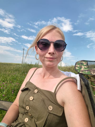 In the city of Kuzminovka, Russia, on a sunny day with a blue sky dotted with white clouds, a woman with blonde hair and sunglasses is captured in a close-up selfie. She is wearing a green sundress with wooden buttons and large pearl hoop earrings. The woman is seated in an outdoor setting, with a field of green grass and purple flowers visible in the background. To her right, a camouflage-patterned bag or chair is partially in view. A telephone pole stands in the distance. The overall impression is one of a casual, outdoor leisure activity.