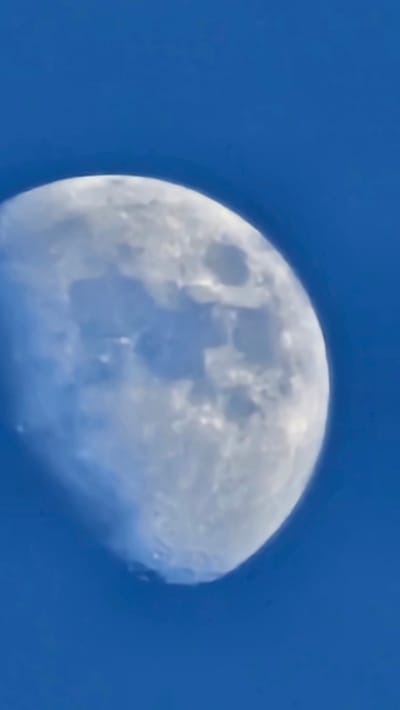 A half-illuminated moon is captured against a clear, bright blue sky. The moon's surface exhibits visible craters and maria, creating a textured appearance. The lighting suggests it is daytime, possibly late afternoon or early morning, when the moon can be seen in the sky. The image is a close-up shot, focusing solely on the celestial body and its backdrop. There is no indication of people, specific activities, or explicit location cues beyond the general setting of the sky. No text is visible in the image.