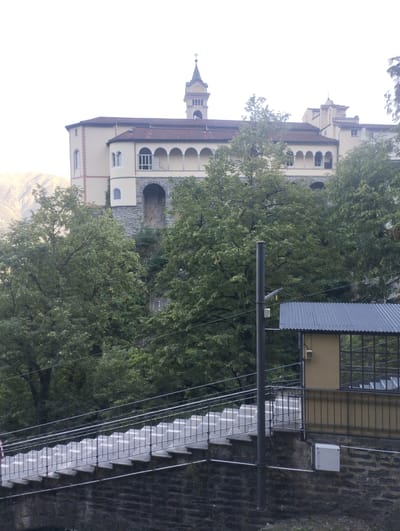 In Orselina, Switzerland, a complex architectural structure dominates the upper portion of the image. This building, likely a monastery or similar religious institution, features a distinctive bell tower with a cross at its apex. The main structure is a light yellow edifice with a red-tiled roof. It has a series of arched windows along its facade, supported by columns. The lower sections of the building are constructed with rough-hewn stone.
In the foreground, a steep, outdoor staircase ascends from the lower left to the middle right of the frame. The stairs are made of concrete, with visible white markers on each step and metal railings on both sides. The staircase is supported by a stone wall. To the right of the staircase, a small, yellow building with a dark, corrugated metal roof is visible. This building has large windows with dark frames. A metal pole, possibly for lighting or power lines, stands to the left of this smaller building.
The scene is framed by abundant green foliage from large trees that obscure much of the lower and mid-sections of the building and the surrounding landscape. The sky is overcast and appears bright white, with no discernible clouds. The overall impression is a blend of natural and built environments.