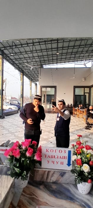 The image displays a semi-covered tiled patio area in Chinabad, Uzbekistan, beneath a prominent metal grid roof structure with several ceiling fans. Two men are central to the foreground. The man on the left, dressed in a dark long-sleeved shirt, dark trousers, and a black-and-white patterned Uzbek skullcap, holds a dark object in his left hand while his right hand is raised near his mouth. The man on the right, wearing a white shirt, a dark blue vest with a name badge, dark trousers, and a similar skullcap, extends his right arm to hold a black smartphone, seemingly taking a selfie or recording video. In the immediate foreground, two decorative white vases hold artificial flowers: one on the left contains pink and white roses, and one on the right contains mixed red, white, and yellow flowers. Positioned between these vases on a patterned marble-like surface is a white sign with "КОГОЗ ТАШЛАШИ УЧУН" (FOR DISPOSAL OF PAPER) written in Uzbek Cyrillic, accompanied by a blue downward arrow. In the background to the left, through a glass partition, parked vehicles (a white car and a dark van) and distant buildings are visible. To the right, inside an open area of a building, several blurry individuals are seated at low wooden tables.