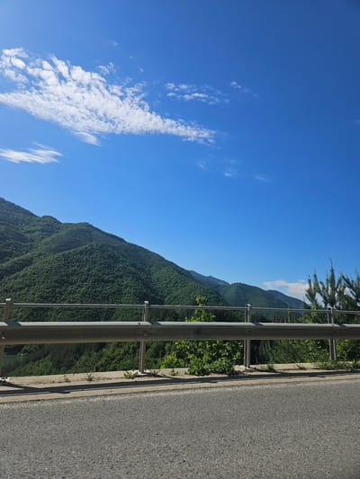 The image shows a view from a road in Liaskovo, Bulgaria. The sky is blue with scattered white clouds. A metal guardrail runs along the right side of the road. Beyond the guardrail, a steep, densely forested mountain slope rises. The road itself is asphalt and takes up the right side of the frame.