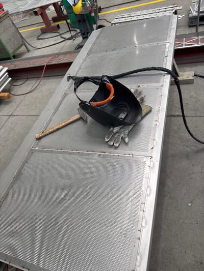 The scene appears to be an industrial setting, possibly in Račice, Czechia, although there is no explicit visual confirmation of the location. An elevated, silver-colored metal structure with a perforated surface dominates the view. Resting on this structure is a welding mask, a pair of work gloves, and a hammer. A thick black cable runs along the top edge of the structure. The environment includes a gray floor and structural supports. A yellow safety line is also visible. Other machinery and materials are partially visible in the background. No people are visible. The scene gives the impression of an inactive workspace with tools temporarily placed on the structure.