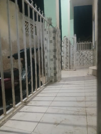 A staircase with metal railings ascends, tiled steps visible beneath. A dark, rectangular opening is visible at the top. To the right, a tiled wall runs parallel to the staircase, with horizontal lines and light-colored tiles. The area below the staircase appears dark and contains some undefined objects. Overall, the image presents a somewhat tilted and narrow view of the staircase and its surrounding environment.
