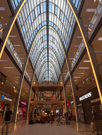 The image depicts the interior of the Ettlinger Tor shopping center in Karlsruhe, Germany. The architecture features a striking, arched glass ceiling supported by metal beams, with integrated yellow lighting illuminating the multi-level structure. The foreground shows a polished floor reflecting the ambient light. On the left, a man in a dark t-shirt and light pants walks towards the viewer. Adjacent to him is a shopfront with a "blue-tomato" logo. Further to the right, the "TALLY WEiJL" store is visible, advertising a sale. In the midground, several people are gathered, some sitting on sofas and chairs in a seating area. More shops and escalators are visible on the upper levels. On the right side of the image, a "SKECHERS" store with its prominent signage is present, with two closed storefronts. In the background, more shoppers are seen dispersed throughout the mall. The overall impression is a well-lit, modern retail environment.