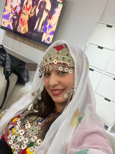 A woman wearing traditional clothing smiles at the camera. She wears a white headscarf, ornate headpiece with dangling silver coins, and a colorful necklace. In the background, a television displays a performance with dancers wearing elaborate costumes. A white cabinet with black handles is visible above. A grey bag with black straps rests on a light-colored surface. The scene is brightly lit, creating a cheerful and festive atmosphere.