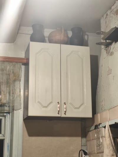 The image depicts an indoor scene, likely a kitchen, with a focus on a light-colored cabinet mounted high on the wall. Three decorative pottery jugs are placed on top of the cabinet, varying in color from dark gray to a light reddish-brown. The cabinet itself has two doors with ornate, recessed paneling and small, floral-patterned handles. To the left of the cabinet, a portion of a window with a sheer, patterned curtain is visible.
The wall to the right of the cabinet appears to be made of roughly textured plaster, with areas of peeling paint revealing a lighter color underneath. A simple metal shelf with two circular cutouts is attached to this wall, and a set of metal tongs hangs from it. Below the cabinet, a wall is visible, painted a neutral, sandy color. Towards the bottom right, a tiled section of the wall is apparent, and a beige towel hangs to the right of the tongs. A black electrical cord with a plug extends from the lower left side of the frame, suggesting the presence of an appliance.
The lighting in the scene is somewhat dim and appears to be artificial, possibly from an overhead light source, casting shadows on the wall below the cabinet. The overall impression is of a rustic or perhaps older kitchen setting. There is no discernible text in the image. The location context provided is Навадворскі сельскі Савет, Belarus.