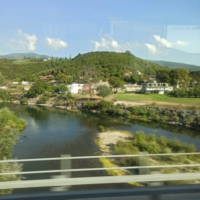 The image captures a scenic view of a river flowing through a rural landscape in the Municipal Unit of Kato Olympos, Greece. A lush green hill, covered in trees and dotted with a few houses, rises behind the riverbank. The river itself is dark and reflects the sky and surrounding greenery. Along the riverbank, there are rocks and vegetation. In the foreground, there are white railings, suggesting the image was taken from a bridge or overpass. The sky is blue with a few scattered white clouds. The overall impression is one of tranquility and natural beauty.
