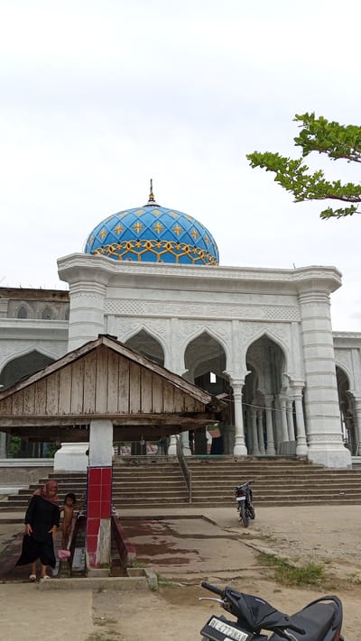 The image depicts a mosque in Beunyot, Indonesia, with a prominent blue and gold dome. In the foreground, a woman in a black hijab and a child stand near a water source with tiled pillars. Several stone steps lead up to the mosque's entrance, which features arched doorways and white columns. A wooden structure with a dilapidated roof is positioned in front of the steps. Two motorcycles are visible, one parked near the steps and another partially visible in the lower right corner. The sky is overcast. The overall scene suggests a public area adjacent to a religious building.