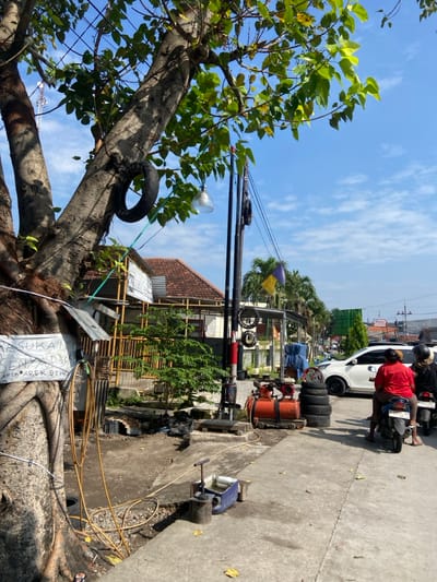 The scene is set in front of a small workshop on the outskirts of a residential area in Kecamatan Tulangan, Indonesia. A large tree trunk dominates the left side of the scene, while a pole with a yellow flag hangs from the top, positioned next to the workshop.  The workshop is a simple structure with a corrugated metal roof and a brick wall.  A sign on the workshop reads "SUKA SENYAP 36" and "AREK AREK DEWI" (spelling and grammar may not be perfect). A man in a red shirt sits on a motorcycle, while another man in a black helmet looks on from the back of another motorcycle.  A white car is parked in the street.  The ground is covered in dirt and debris.  The sky is blue and cloudless.