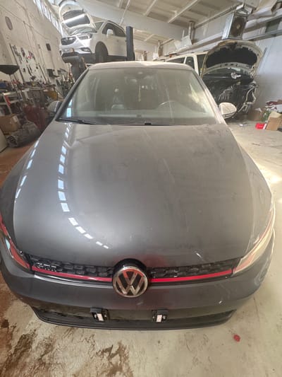 In an auto repair shop in Schkopau, Germany, a dark gray Volkswagen GTI is parked in the foreground. The car's hood is raised, showing a dusty surface. The background is filled with other vehicles and repair equipment. To the left, a white Opel Mokka is on a lift with its hood open. Further back, a white SUV has its hood propped open, revealing its engine. The shop appears cluttered with tools, boxes, and car parts, suggesting ongoing repair work. The lighting is dim, with fluorescent lights visible on the ceiling.