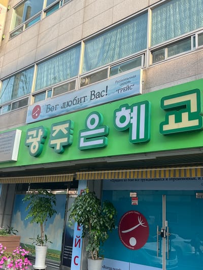 The image shows a street-level view of a building facade in Gwangju, South Korea. A prominent green sign features large, illuminated Korean characters that read "광주 은혜 교회" (Gwangju Grace Church). Above this, a smaller blue and white sign displays Russian text: "Русскоязычная церковь «Грейс»" (Russian-speaking Church "Grace") and "Бог любит Вас!" (God loves you!). The building has several windows with sheer curtains. Below the green sign, a glass door is visible with yellow and gray striped awnings above it. There are two potted trees in front of the glass door, one smaller and one larger. A partially visible advertisement or poster with blue and white imagery is also present on the left side of the door. A CCTV sticker is affixed to the door. The overall impression is that of a religious institution or community center in an urban setting.