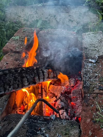 The image shows a close-up view of a fire burning in a makeshift brick fire pit. The fire is fueled by logs and embers, with visible flames and smoke rising. There is a metal poker resting in the fire pit. The surroundings include grass and what appears to be a concrete structure in the background. The scene is outdoors, likely in a rural or suburban setting, given the presence of vegetation and the informal construction of the fire pit. No people are visible in the image. The location is described as being in Imelevo, Russia.