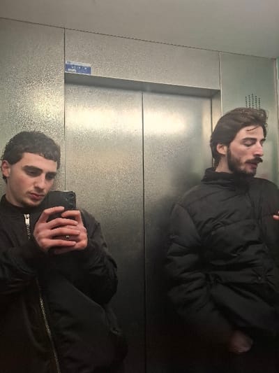 A person in a black jacket is seen reflected in a metallic surface, likely a mirror or elevator door. The reflection shows the person holding a phone, presumably taking a photo of themself. The background is a plain, metallic surface with horizontal lines. The lighting is diffused, creating a soft reflection. The composition of the image is centered on the reflection.