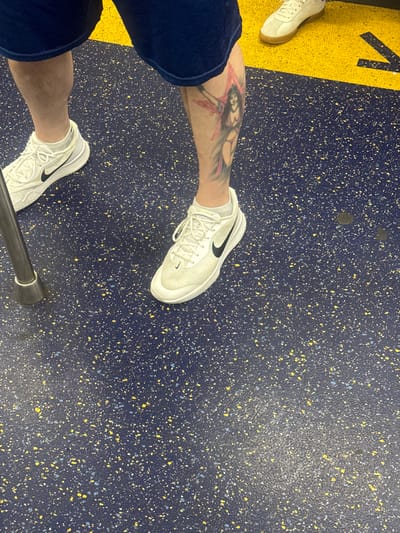 The image shows a close-up view of a person's legs and feet standing on a speckled blue and yellow floor, possibly in a public transportation setting. The person is wearing dark blue shorts and white Nike sneakers. A tattoo of a woman with a star is visible on the person's left calf. A silver pole is also present in the lower left corner of the image. The floor has a pattern of small yellow and white flecks embedded in it, with a yellow arrow pointing to the right in the upper right portion of the image. The overall lighting suggests an indoor environment. No specific landmarks or signs indicating the city of Hoboken, United States of America are visible in the image.