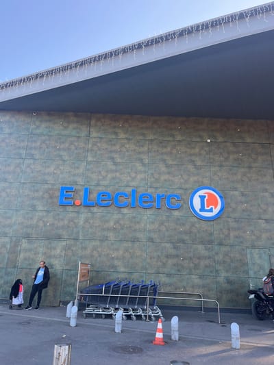 The image shows the exterior of a large, modern building in Saint-Brice-Courcelles, France, identified by the coordinates provided. The building is a E.Leclerc supermarket, as evidenced by the large blue sign with the E.Leclerc name and the red and blue "L" logo. The building facade is constructed of textured, gray, paneled material. A white awning extends over the front entrance. A row of eight shopping carts are parked in front of the entrance. A man in a blue shirt and jeans stands near the entrance, and a child in a black jacket is standing near a shopping cart in front of him. Another man in a black jacket is standing beside a motorcycle with a license plate visible. The ground is paved with gray asphalt. There are several orange and white traffic cones in front of the building. The sky is clear and blue.