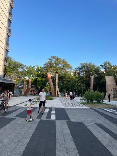 In Ivanovskaya, Russia, a wide paved walkway is populated by several individuals and groups. On the left, a person rides a bicycle, wearing sunglasses and a light-colored t-shirt. Behind them, a group of people walk. In the foreground, a young boy on a scooter with red shorts and a white t-shirt is moving across a crosswalk marked with dark and light grey stripes. To his right, a man in a white t-shirt and black shorts walks. In the background, more people are strolling on the walkway, and a few are visible on bicycles. The environment features modern wooden sculptures, lush green trees, and a tall building on the left. The sky is a clear, bright blue.