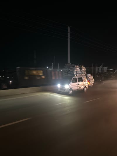 In Mowe, Nigeria, a white van overloaded with sacks of goods drives down a highway at night. The van's headlights illuminate the road, and its taillights are visible. The sacks are stacked high on the roof and secured with netting. The van is in motion, indicated by the motion blur of the surrounding environment and the streaks of light from the headlights. The highway appears to be elevated, with a concrete barrier on the left side. In the background, there are some blurred structures and power lines against a dark night sky.