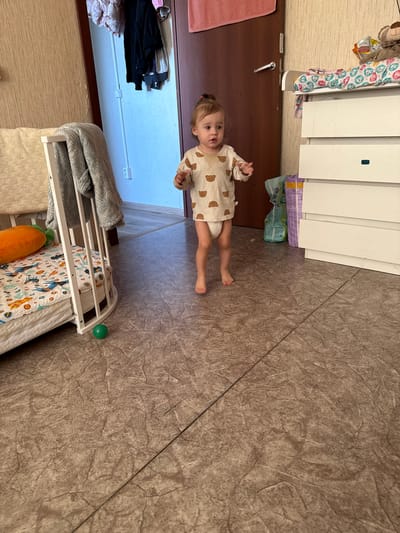 In the image, a toddler stands in the center of a room with a tiled floor. The toddler is wearing a light-colored bodysuit with a teddy bear pattern and a diaper. The toddler has a small amount of food on their chin. To the left of the toddler is a white crib with a grey blanket draped over it. Inside the crib, there is a mattress and an orange pillow shaped like a carrot. On the floor in front of the crib is a small green ball. To the right of the toddler is a white dresser with several drawers. On top of the dresser is a stuffed bear and some baby blankets. Behind the dresser, there are packs of diapers. In the background, a closed door with a silver handle is visible, and a section of a closet with hanging clothes can be seen to the left of the door. The walls appear to be covered in a textured wallpaper. The lighting suggests it is daytime. The scene appears to be an indoor nursery or child's room, likely in a residential setting in Vylygort, Russia, given the provided context.