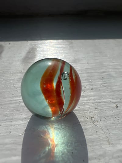 This close-up, high-angle shot features a single, clear glass marble with swirling bands of orange and white. The marble is positioned on a textured, light-colored surface that appears to be wood, possibly painted and weathered, with visible grain and small imperfections. A dark, vertical object, likely a post or wall, is partially visible on the left side of the frame, casting a shadow. Sunlight illuminates the scene, creating a distinct shadow of the marble to its right, which refracts light into a spectrum of colors, hinting at a sunny day. A small air bubble is captured within the marble. The overall impression is one of stillness and a focus on the play of light and color on a simple object. The setting suggests an outdoor environment in the United States, given the provided location context. There is no visible text in the image.