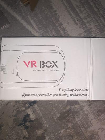 The image shows a white cardboard box with the text "VR BOX" and "VIRTUAL REALITY GLASSES" printed on it. Below this text, there is a quote: "Everything is possible if you change another eyes looking to this world." The box is placed on a gray, textured surface, likely a floor or carpet. There are no people or visible actions taking place in the image. The lighting suggests an indoor environment. The provided context of "Vasilievka, Kyrgyzstan" does not appear to be directly related to the visual content of the image itself, as there are no geographical indicators present.