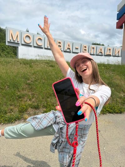 The image shows a young woman with long brown hair, wearing a pink baseball cap, a white t-shirt, a plaid shirt tied around her waist, and light green sweatpants. She is holding a pink-cased smartphone in her right hand, which is outstretched towards the camera. Her left arm is raised, with her hand waving. She has bright pink and blue fingernail polish. The background features a large sign with Cyrillic lettering that reads "МОСКВА-ВЛАДИ", indicating the location is likely near Moscow, Russia. The sign is set against a backdrop of a cloudy sky and a grassy slope with some scattered yellow flowers. The woman is positioned on a paved surface, possibly a road or pathway, with a grassy embankment behind her.