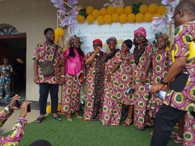 The image depicts a group of individuals standing in front of a decorated backdrop. The backdrop features a sign that reads "First Baptist Church Teenage Department" and is adorned with yellow balloons and purple and white flowers. The individuals are all dressed in colorful, patterned clothing. Two males are standing to the left of the group, with one wearing a green and black shirt and the other a gray shirt. In the center of the group, a woman in a pink and green patterned dress wipes her eye with a tissue. Behind her stand four women, also wearing similar pink and green dresses. On the right side of the group, a man in a black shirt and patterned pants, is looking directly at the camera. In the background, a young boy can be seen wearing a patterned shirt and shorts. The image was captured in front of the First Baptist Church Teenage Department in Yenagoa, Nigeria, located at 4.932 latitude and 6.268 longitude. The scene is likely a gathering for a church event, potentially a celebration or a special occasion.