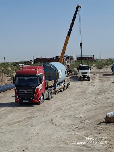 In an outdoor, dusty, construction-like environment under a clear sky, a large yellow crane is positioned with its boom extended upwards and a hook hanging. In the foreground, a red semi-truck with a white and red stripe design is parked. It has a "SANY" logo visible on the windshield and on the side of the truck. The truck is towing a large, blue, cylindrical container on a flatbed trailer. To the right, a white truck is visible with people on top. In the background, there are several small, dark-colored buildings with red roofs and some light-colored structures with blue roofs. There are also some sparse, green, bushy plants and trees. The ground is dirt and sand, with tire tracks visible. A timestamp "Galaxy A56 5G 22 июня 2026 г. 16:18" is present in the bottom right corner. This appears to be an industrial or construction site in an arid region.