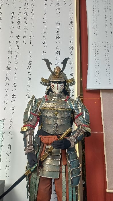 The image displays a detailed, full-body suit of traditional Japanese Samurai armor, presented on a mannequin in a static, upright pose within what appears to be a cultural exhibition space or museum in the Special Capital Region of Jakarta, Indonesia. The armor, predominantly in metallic silver/grey and teal-green hues, features gold accents and red lacing. Key components include a multi-layered helmet (kabuto) with two prominent black fin-like crests and a golden circular ornament, and a distinctive white facial mask (mempo) with red markings resembling a fox or demon. The torso armor (dou) consists of riveted metallic plates. Layered, teal-green metallic plates form the shoulder guards (sode) and thigh guards (haidate), while the arm guards (kote) are segmented. The mannequin wears reddish-brown trousers beneath the armor and black gloves, with an orange/rust-colored fabric sash (obi) tied at the waist. A short sword (wakizashi) with a golden, textured scabbard is held in the mannequin's right hand, and the hilt and part of the scabbard of a long sword (katana) are visible at its left side. The background features a large vertical white panel on the left adorned with black Japanese calligraphy, and a vertical red-brown panel on the right, partially obscured by a smaller white scroll also featuring Japanese text. The scene is evenly lit, suggesting controlled indoor lighting typical of a display environment.